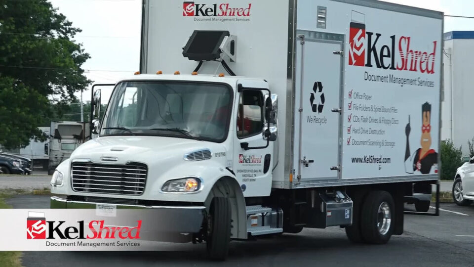 A white KelShred truck, branded with "Document Management Services" and recycling symbols, drives on a road. Featuring a cartoon superhero character, the truck highlights KelShred's secure shredding services. The prominent logo assures customers of their commitment to confidential waste disposal.