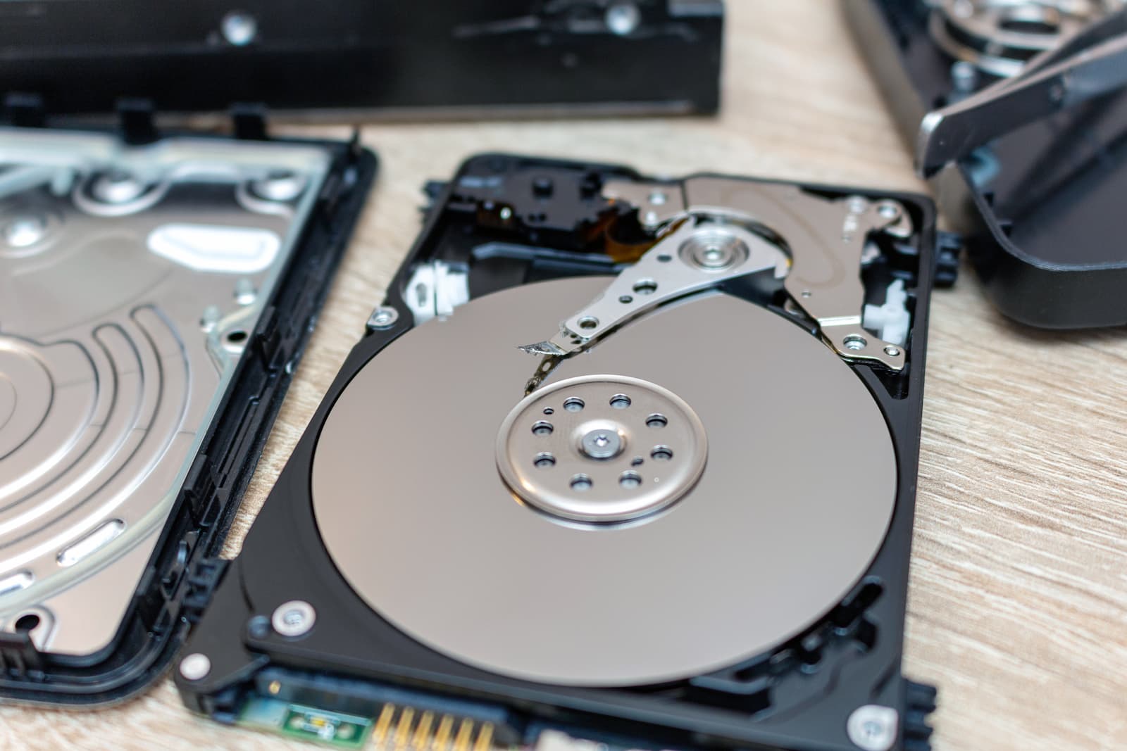 Open hard disk drives rest on a wooden surface, their intricate platters and internal components exposed. One drive is fully open, while another is partially disassembled in the background, reminiscent of meticulous document shredding processes in confidential waste disposal.