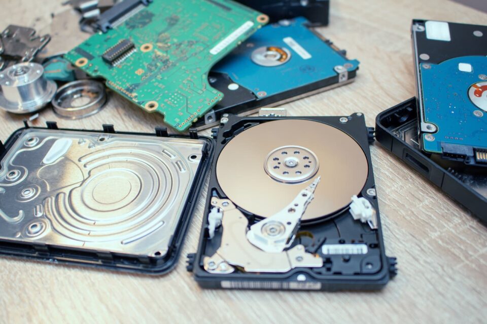 Disassembled hard drives with visible internal components, including platters, read/write heads, and circuit boards. The parts are spread out on a wooden surface, showcasing the intricate details of the hardware prior to secure shredding services ensuring confidential waste disposal.