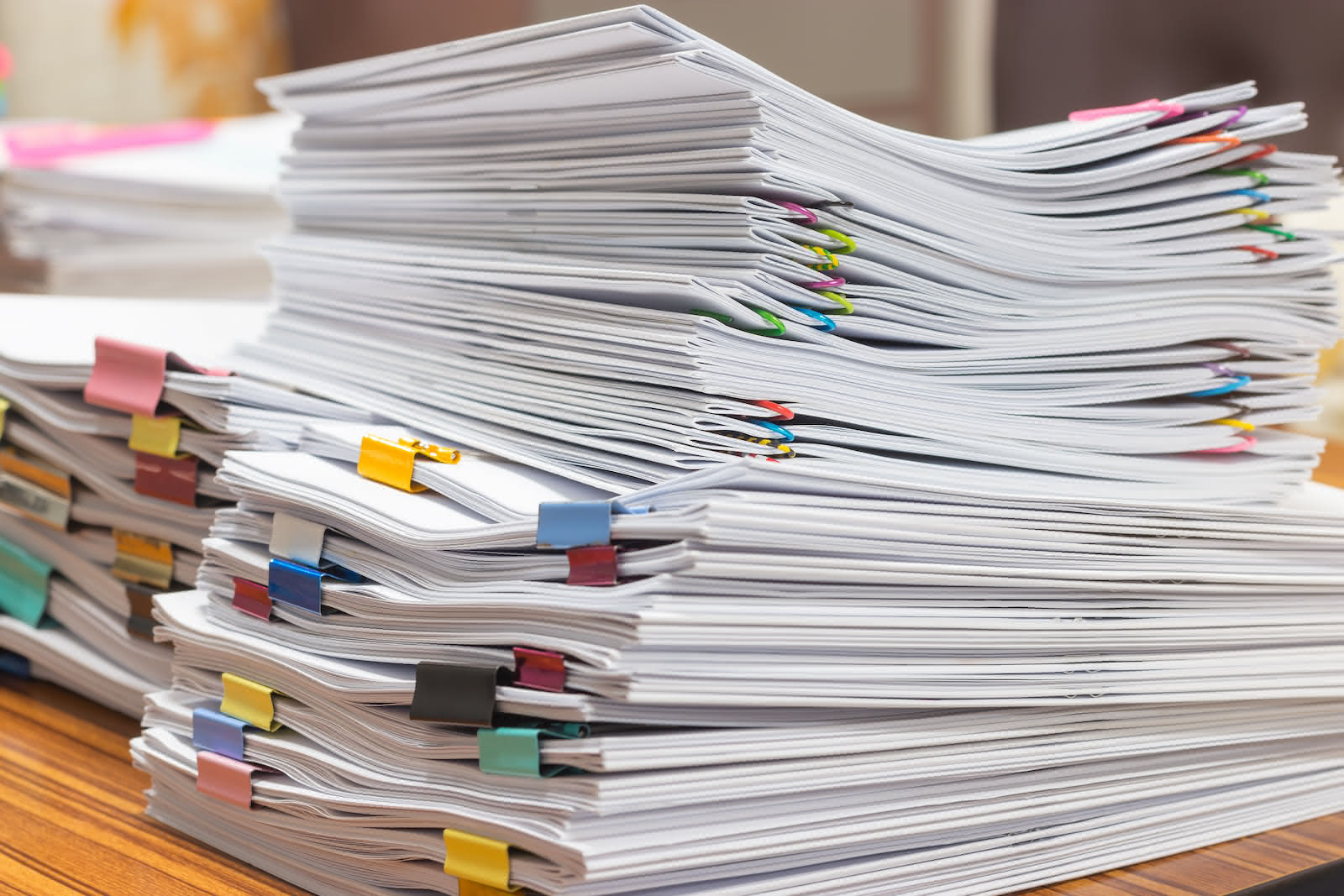 A large stack of documents is neatly arranged on a wooden surface. Bound with various colored binder clips, including pink, blue, yellow, and black, these papers await secure shredding services. Some stacks are taller than others, indicating different volumes of paperwork.