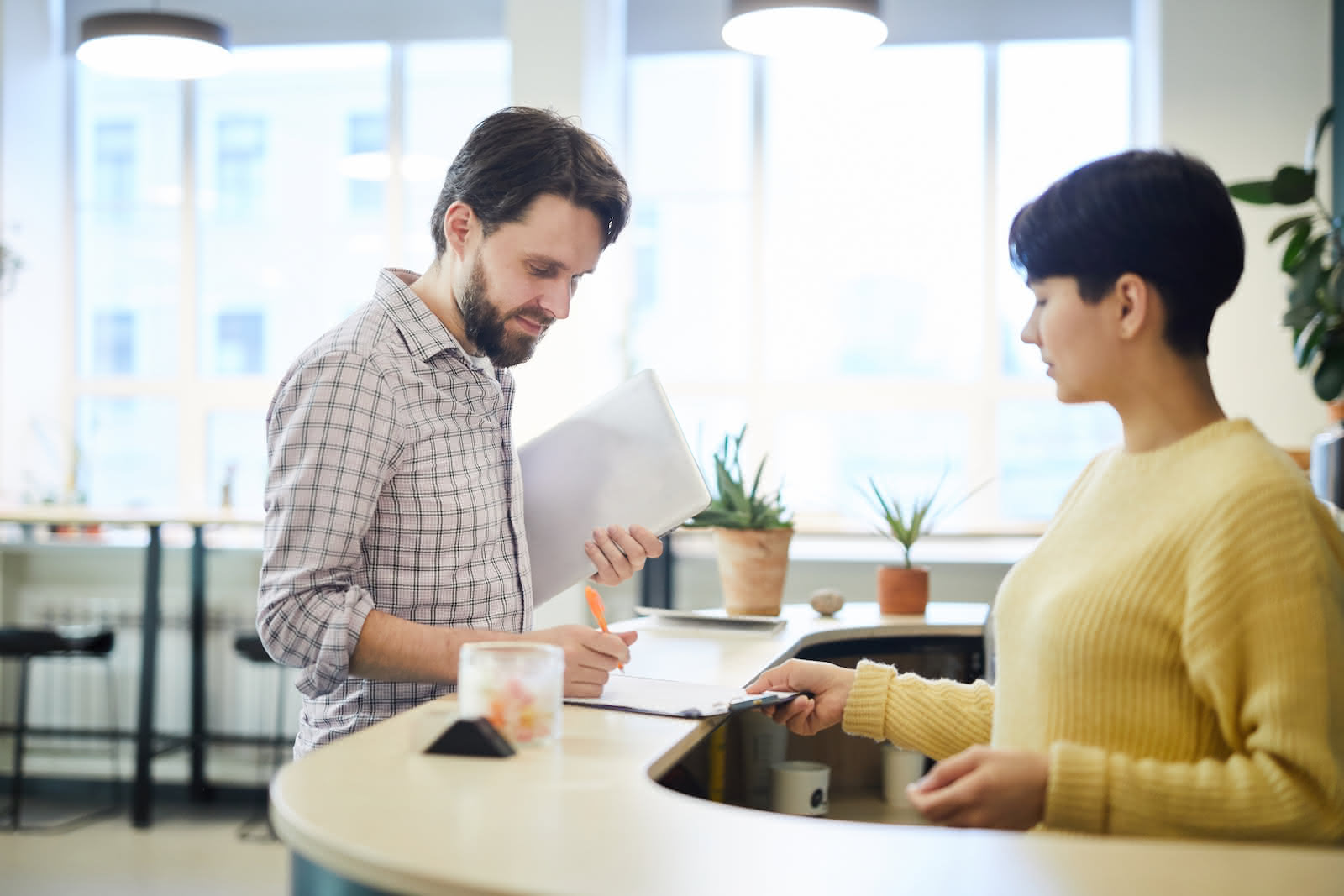 A man is standing at a reception desk, signing a document held by a woman. The desk has a small potted plant and a container of candy on it. Both individuals are in a well-lit room with large windows in the background, ensuring they can discuss secure shredding services confidently.