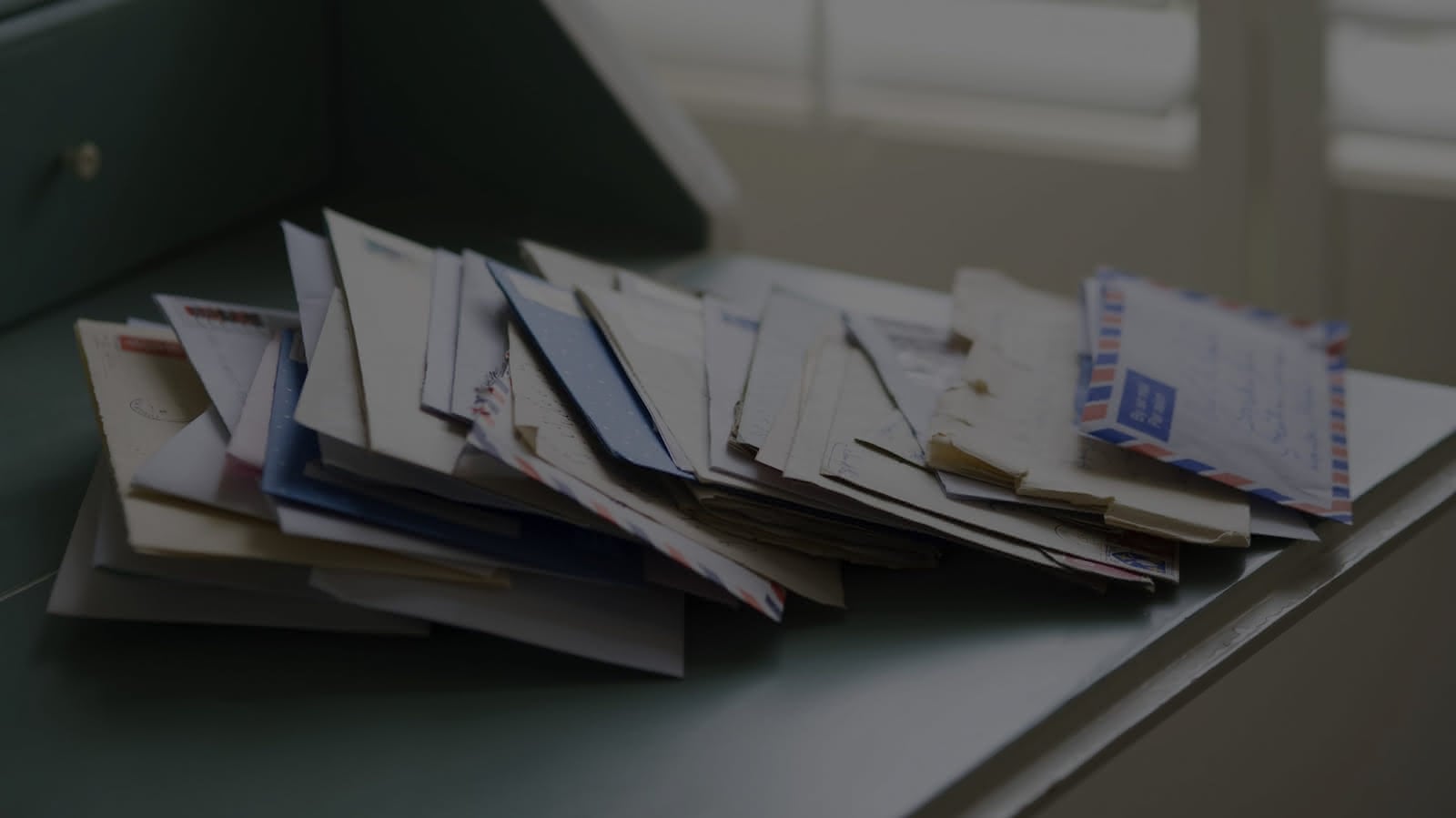 A stack of various envelopes and letters is scattered on a gray surface, some bearing airmail markings and others plain. The background includes blurred, vertically-slatted window blinds allowing diffused light into the room, suggesting the need for confidential waste disposal to protect sensitive information.