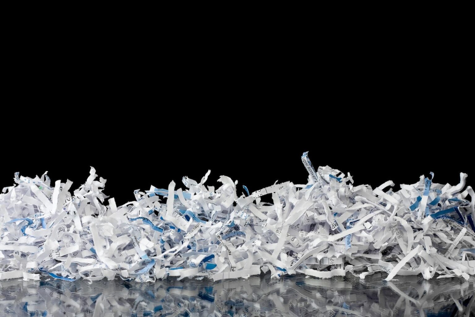 A pile of shredded paper from secure shredding services, with interspersed blue and white strips, lies against a solid black background. The shreds reflect slightly on a glossy surface below them, creating a textured and chaotic appearance.