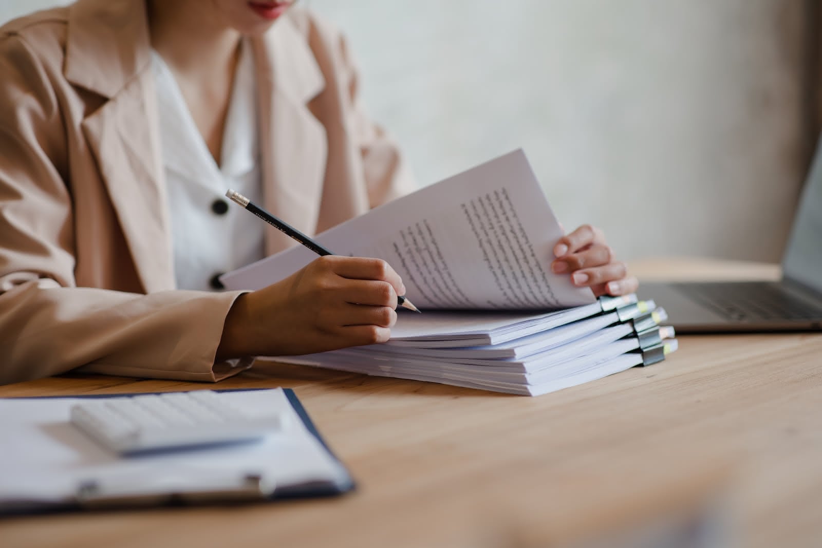 A person is sitting at a desk with a stack of papers. Holding a pencil, they appear to be reviewing or marking the documents. A clipboard with papers and a calculator are also on the desk, and a laptop is visible in the background. The scene hints at the importance of confidential waste disposal after document shredding.