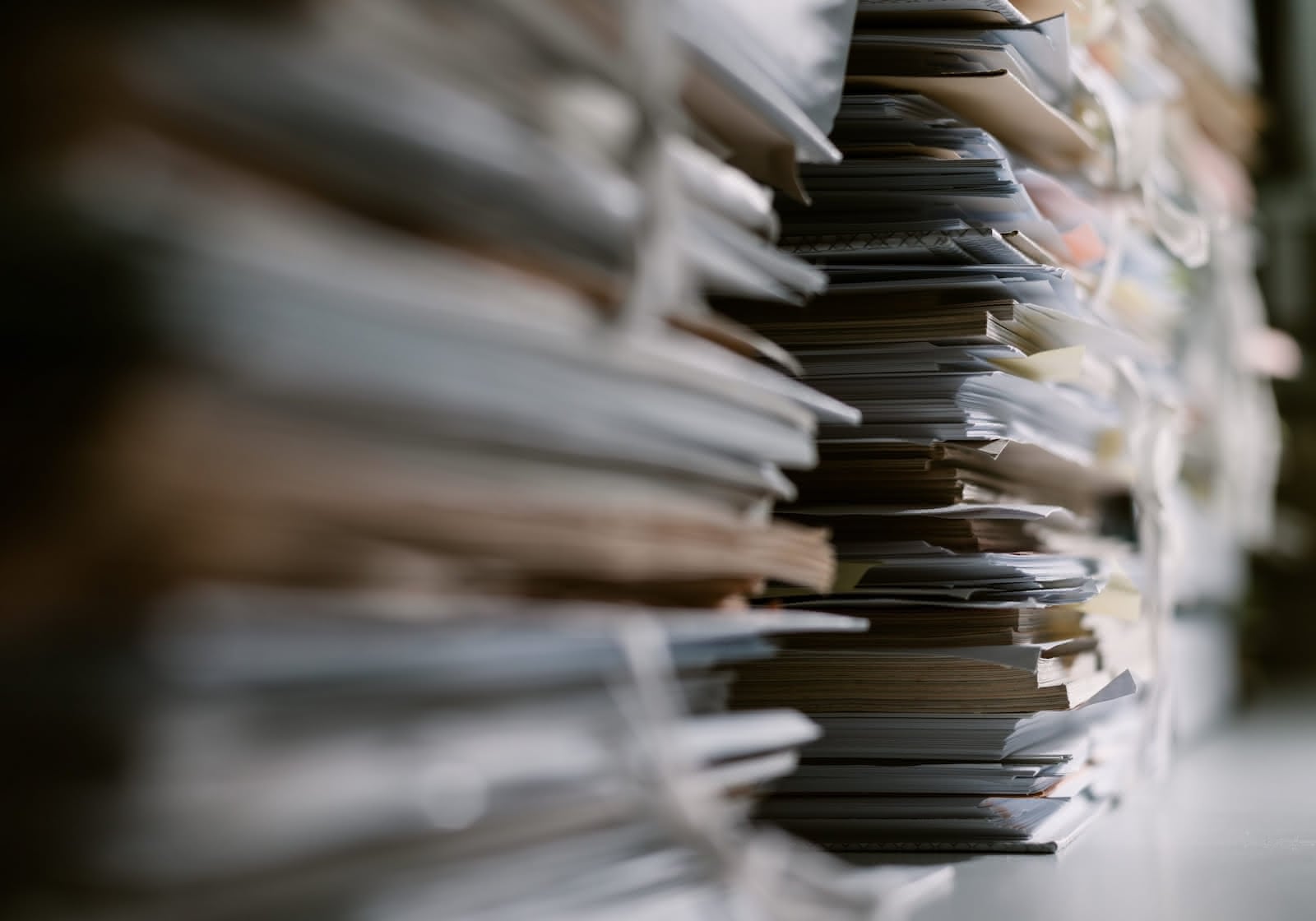 Close-up of a large stack of papers and documents piled up haphazardly. The focus is on the edges of the papers showing varying thicknesses and textures, begging for document shredding. The background is blurred, emphasizing the volume and disorganization of the stack.